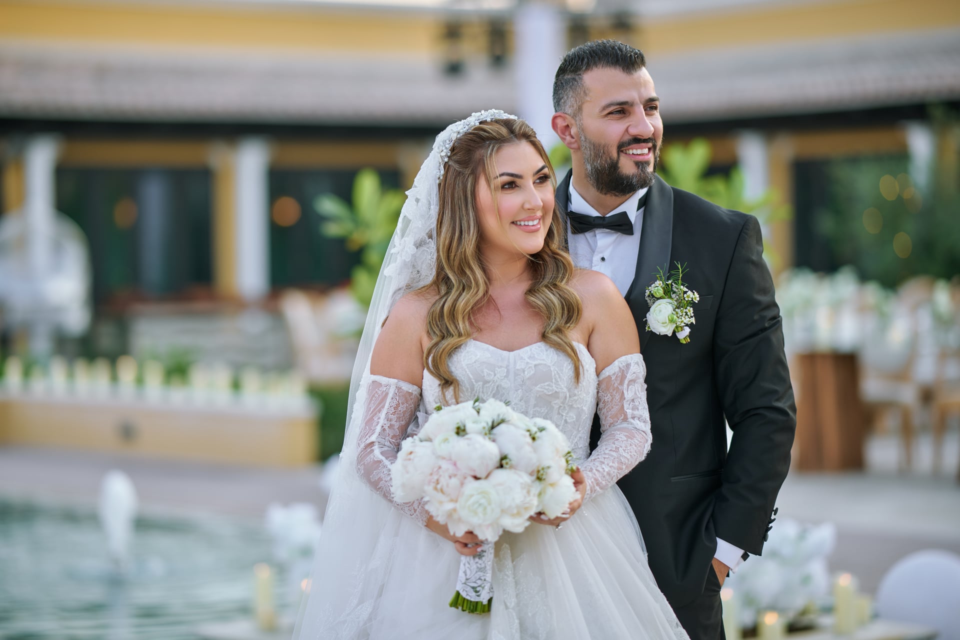 Bride and groom beside a fountain, holding an ivory peony bouquet.
