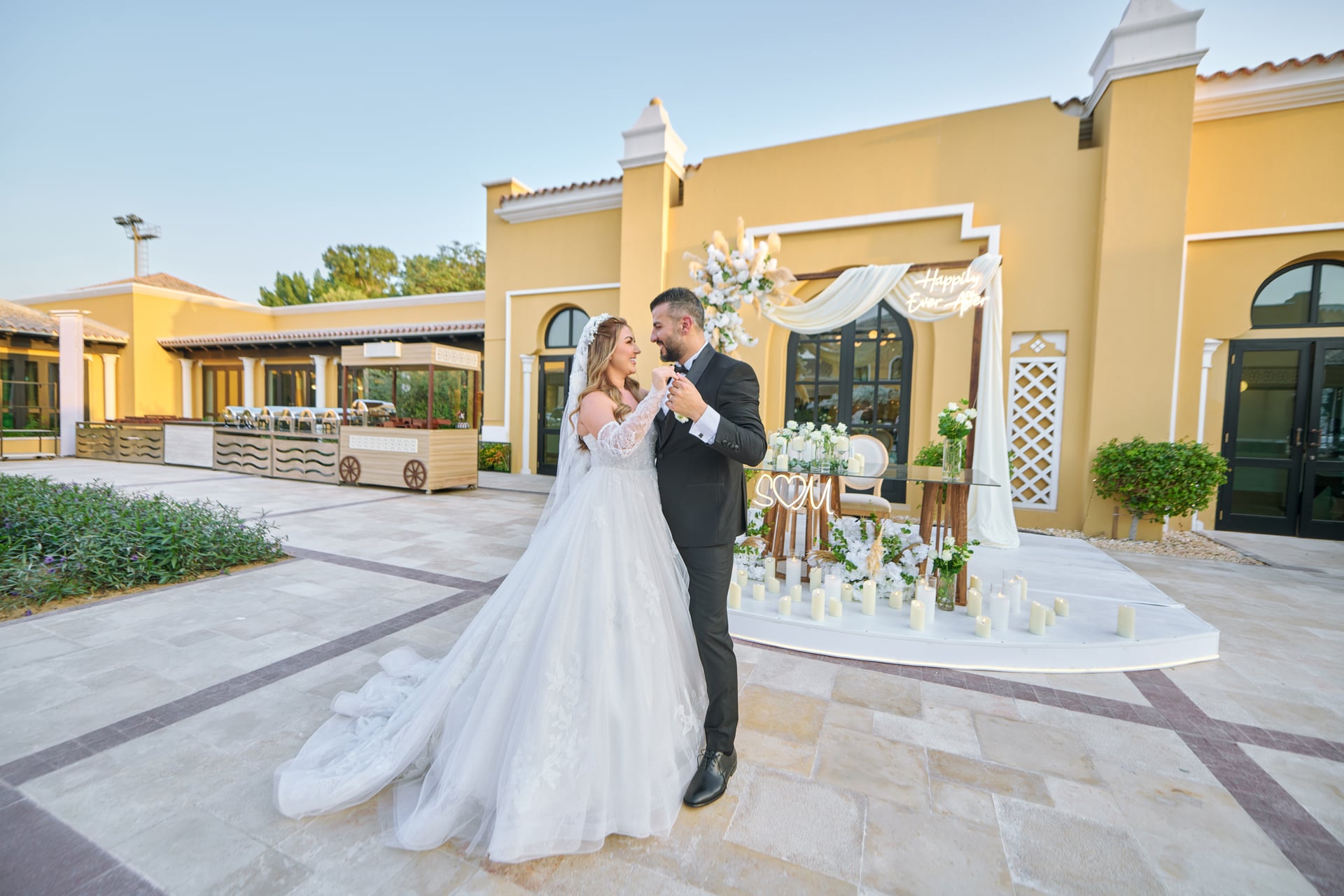 Bride and groom beside a white floral wedding setting in a sunlit courtyard.